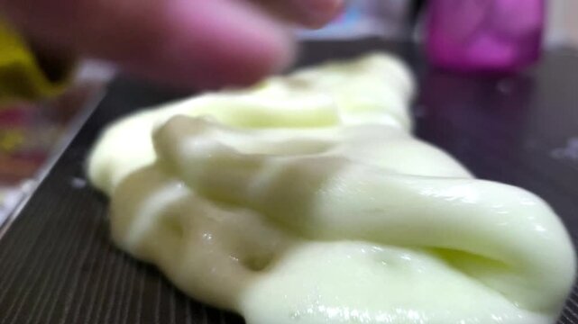 Close-up of child's hands stretching and pulling soft pale yellow fluffy slime on a dark wooden table, fine motor skills activity and ASMR