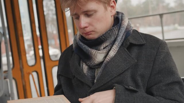 White creator checking notes on tram seat, scarf and beret, pensive young man writing ideas in notebook beside rainspeckled window, vintage interior and waterfront passing by, muted overcast mood