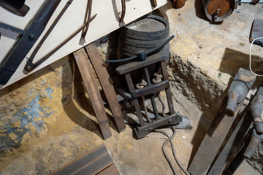 An old-fashioned screw press made of wood and rope sits next to small barrels in a rustic, earthen-walled cellar or workshop area.