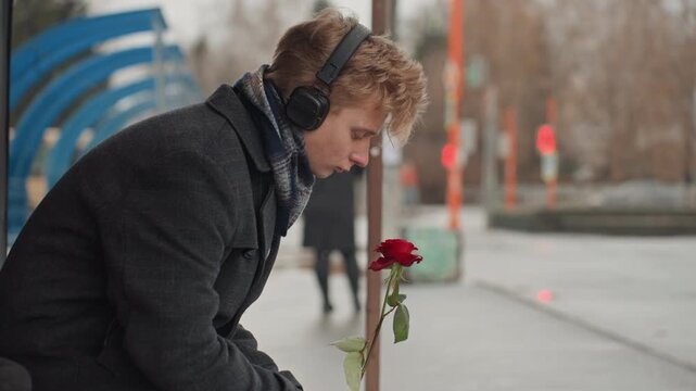 White creator offering single red rose at bus platform, headphones on, winter coat and scarf, urban sidewalk scene gentle hopeful gesture, candid mood, commuter backdrop and soft overcast lighting