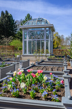 Glass pavilion greenhouse surrounded by blooming garden beds in sunny weather.