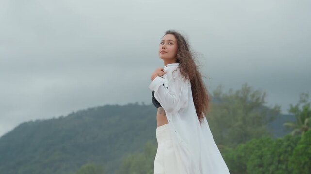 Woman overlooking island hills under clouds, flowing white coverup, windblown hair, tropical trees and ridgeline, dramatic sky, contemplative stance, solitude mood, nature retreat atmosphere