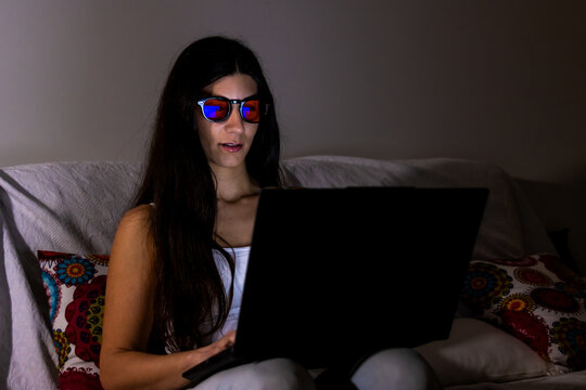 Woman wearing blue light blocking glasses working on laptop in dark room