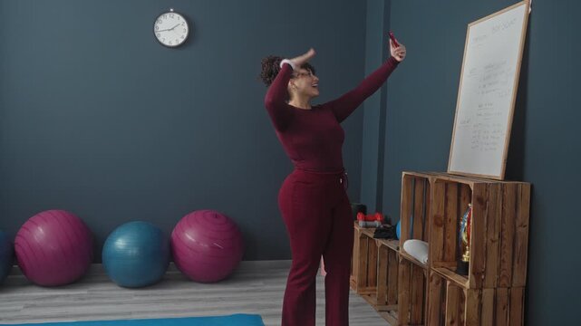 Woman holds smartphone at arm's length for selfie, touches hair beside exercise balls and wooden crate shelf in gym studio; self care confidence.