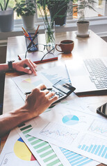 Person working on financial documents with calculator and charts. Hands focus on data analysis on wooden desk. Natural light from window illuminates workspace. Ideal for business, finance