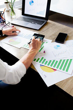 Person analyzes colorful charts on a wooden desk. Their hands move between papers and a smartphone. Laptop displays a pie chart nearby. Focus on data interpretation and professional work. Ideal for fi