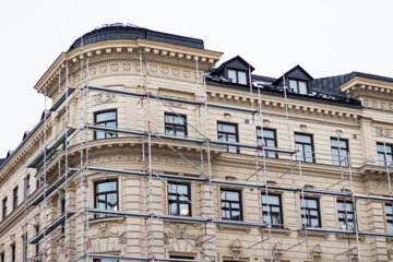 Renovation scaffolding surrounding historic building with ornate architecture and multiple windows, showcasing detailed cornices and a sloped roof in an urban setting © Yahor
