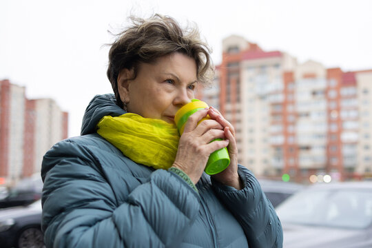 Mature woman in warm jacket and bright scarf holding reusable coffee cup on urban balcony, thoughtful look, city background.
