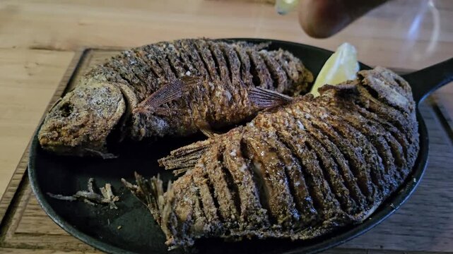 Two slightly overcooked fried crucian carp on black pan with fresh lemon being squeezed over crispy fish in rustic homemade cooking kitchen atmosphere