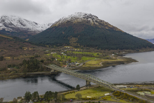 Aerial view of Ballachulish Bridge spanning Loch Leven with snow-capped mountains and lush green hills under an overcast sky in Ballachulish, Scotland, United Kingdom.