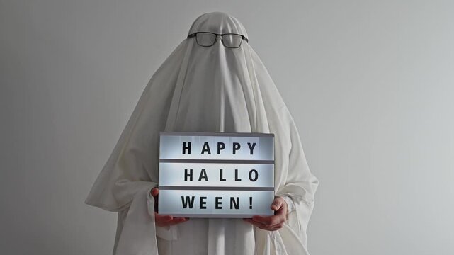 man dressed as ghost holds happy halloween neon sign, october 31