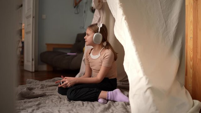 Young white girl sitting on rug wearing headphones and holding controller inside cozy blanket fort, afternoon sunlight, focused expression, casual socks, .