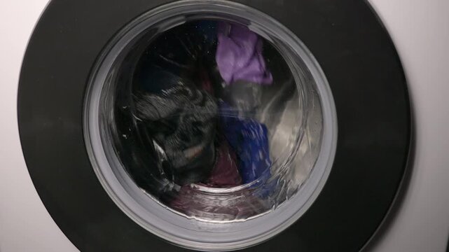 Washing machine drum spinning clothes during cycle. Close-up view of a washing machine drum spinning with colorful clothes and water during a laundry cycle, representing household chores