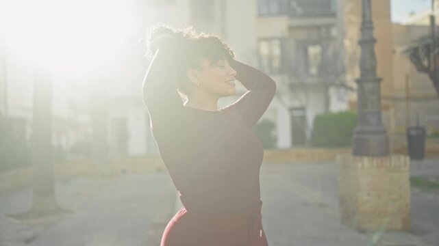 Woman adjusts curly hair with hands, smiling face, backlit by sun near lamppost on street; sunlit serenity.