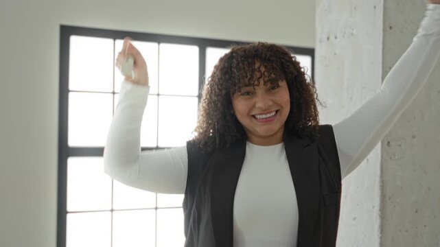 Woman with curly brunette hair holds keys high with hand raised, smiling in empty building interior by window and concrete pillar; new home joy.
