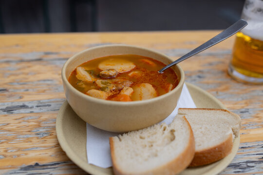 Fresh goulash with beef and vegetables in a ceramic bowl. Sliced bread rests beside the bowl. Beer glass sits nearby on a rustic wooden table.