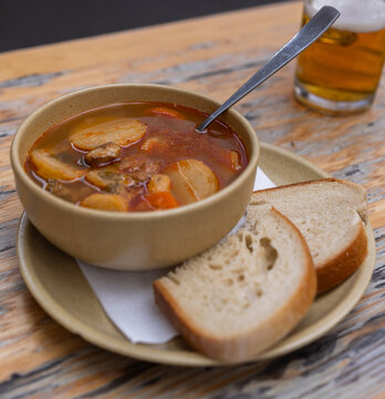 Warm goulash with meat, vegetables, bread, and beer in background. Meal is presented on a rustic wood table.