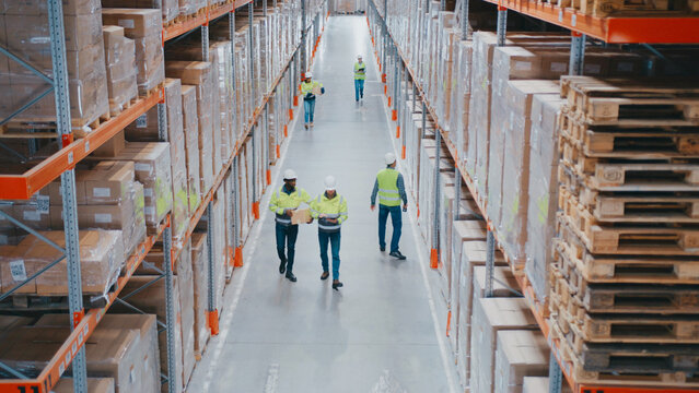 Hardworking warehouse employees wearing safety helmets and vests walking between high pallet racks. Men scanning items with devices. Workers transporting boxes or checking inventory data on tablet.