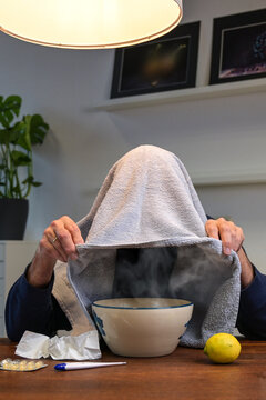 Home remedy against cold and flu, man doing steam inhalation over a bowl with hot herbal water with a towel over his head at a table, health concept, vertical