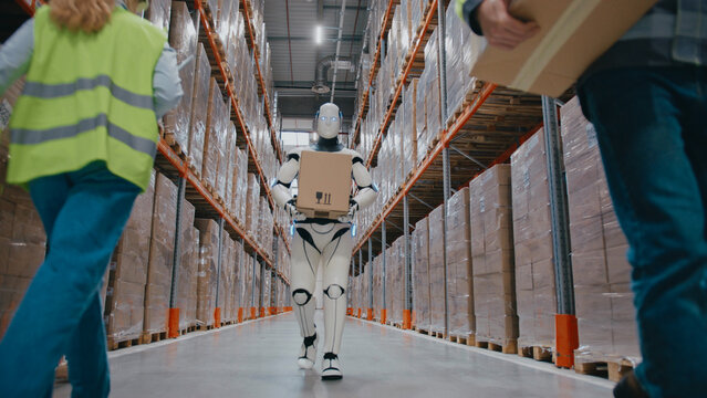 Warehouse robot carrying parcel through aisle while two female workers in safety vests scanning boxes on shelves. Another employee walking past with package during busy automated fulfillment process.