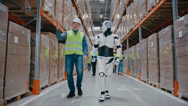 Supervisor in reflective vest pointing upward while walking with humanoid robot through warehouse aisle. Two workers in safety gear moving past. Carrying box and checking tablet during active workday.