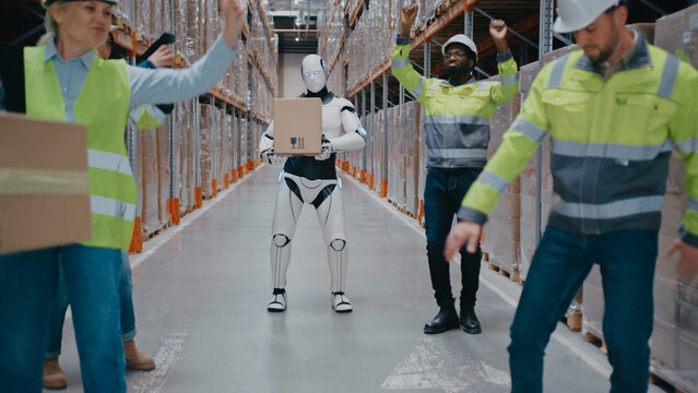 Humanoid robot holding package while walking through warehouse aisle surrounded by cheerful workers. Team raising arms and celebrating successful cooperation between automation and human staff.