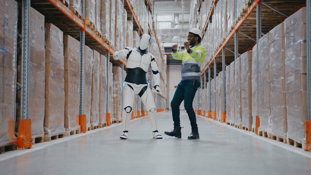 Energetic African American male in safety uniform dancing with humanoid robot inside warehouse. Celebrating teamwork or enjoying moment during automated logistics collaboration. Having fun together.
