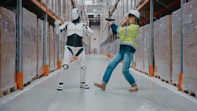 Playful female warehouse worker in helmet holding scanner while posing next to robot. Engaging in fun moment or filming content together inside automated storage facility surrounded by packages.