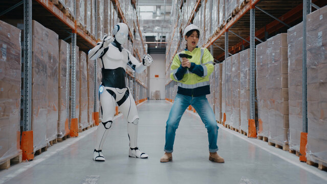 Female warehouse worker playfully pointing barcode scanner at humanoid robot pretending to block in fun pose. Both standing between high shelves stacked with boxes in automated logistics facility.