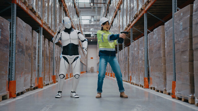 Humanoid robot and female warehouse worker dancing together between tall racks filled with boxes. Woman holding barcode scanner while robot poses with arms wide in funny moment at distribution center.
