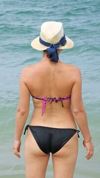 Woman Enjoying Peaceful Seaside Walk. Relaxed Woman In Hat Experiencing Tranquil Oceanfront Journey. Woman In Stylish Sunhat And Bikini Strolling Along Calm Coastal Waves With Peaceful Serenity