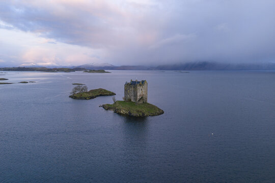 Aerial view of Castle Stalker on a small island in Loch Laich with snow-capped mountains and soft twilight clouds Appin, Scotland, United Kingdom.
