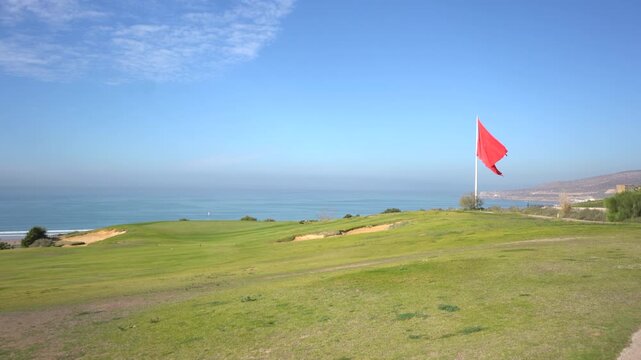 Wide shot of a coastal golf course in Morocco with the national flag waving in the wind against a blue ocean backdrop. Ideal B-roll for luxury travel, tourism, and golf vacation projects.