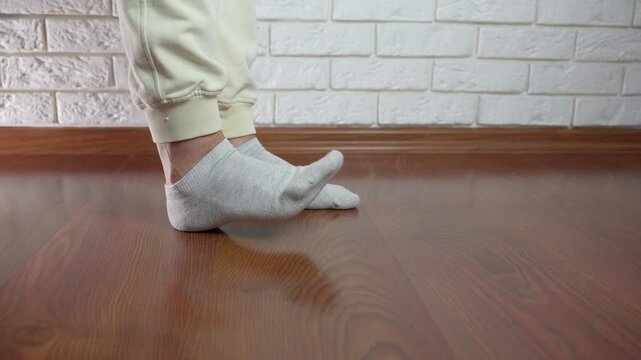 Person massaging painful foot with ankle pain. Woman in sweatpants standing on a wooden floor, massaging her sore foot and ankle due to pain from an injury or cramp, experiencing discomfort