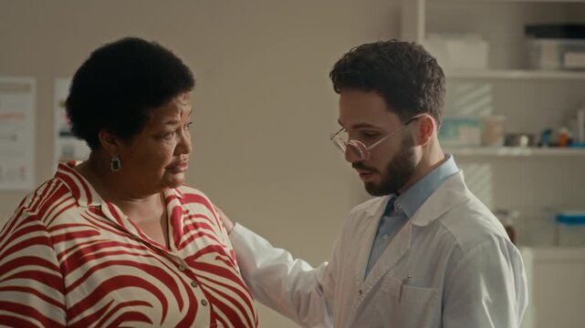 Medium shot of Middle Eastern male doctor using digital tablet to explain medical information to senior Black female patient during healthcare appointment in modern clinic