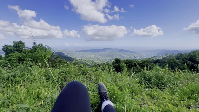 A POV scene shows legs resting in tall green grass while overlooking a wide tropical mountain valley. Rolling hills, lush vegetation, white clouds, and a vivid blue sky create a bright, peaceful outdo
