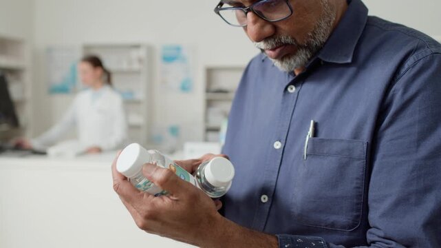 Side view shot of senior Indian man holding bottles of pills in hand and reading labels while choosing medication in pharmacy, copy space