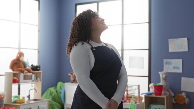 Woman teacher in dark apron points to toys and gestures with smiling hands inside a brightly lit kindergarten building; nurturing joy.