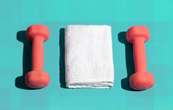 Two pink dumbbells and white towel on blue background, fitness equipment for exercise and healthy lifestyle, top view with copy space.