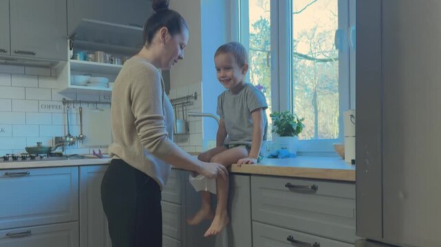Mother talks to child sitting on kitchen counter encouraging him to blow nose. Everyday parenting care and family routine scene.