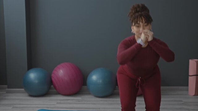Woman squats with hands clasped near face and knees bent on blue yoga mat in studio; wellness focus strength.