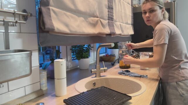 Woman tidies kitchen placing kettle and organizing countertop. Everyday home cleaning routine and domestic order maintenance scene.