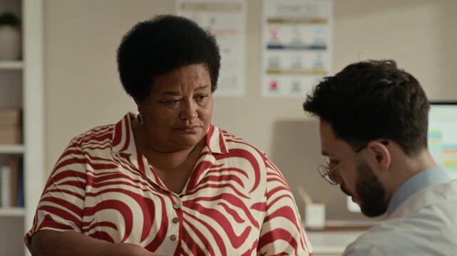 Tilt up of Middle Eastern male doctor measuring blood oxygen levels on senior Black female patient using finger pulse oximeter in clinic office