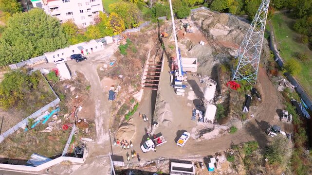Vertical aerial drone footage of an active construction site where a drilling rig and construction crew perform foundation and ground engineering work. The scene highlights excavation, heavy machinery