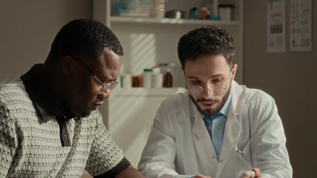 Tilt up of Middle Eastern male physician showing Black male patient how to operate glucometer during diabetes consultation in bright clinic office