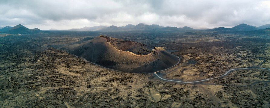 Aerial view of a massive volcanic crater surrounded by barren lava fields and a winding road under a cloudy sky Lanzarote, Canarias, Spain.