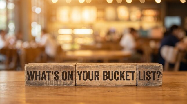 Wooden blocks spell out What's on your bucket list? sitting on a wooden table, with a blurred coffee shop in the background. Evoking thoughts of travel, life goals and adventure. 