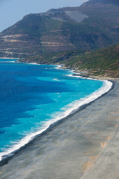Aerial view of Plage de Nonza featuring dark grey pebbles and turquoise Mediterranean waters along the rugged mountains of Corse, France.