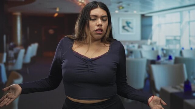 Woman hispanic curvy shrugs with open palms and puzzled expression in restaurant dining building with visible tables and chairs; confusion.