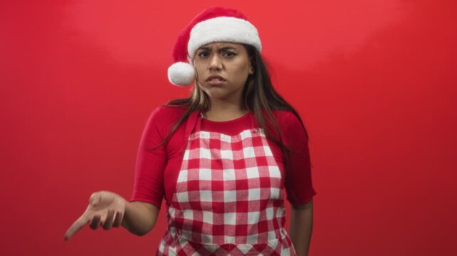 Woman hispanic plus size wearing santa hat and red checked apron points finger to lips and gestures palm up in studio; annoyance.